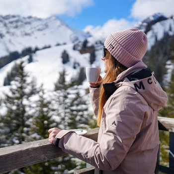 Frau mit Kaffee auf einer Terrasse in Brand mit verschneiten Bergen im Hintergrund in Vorarlberg.