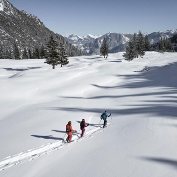 Drei Wanderer mit Schneeschuhen in einer verschneiten Winterlandschaft mit Blick auf alpine Gipfel unter blauem Himmel.